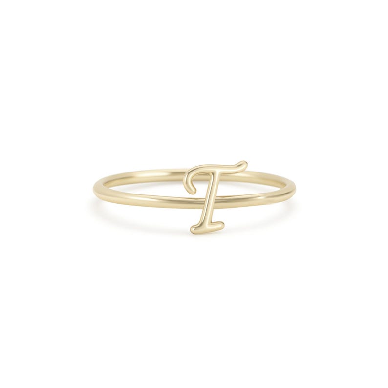 May include: A gold-coloured ring with the letter "T" in a delicate, cursive font. The ring has a thin band and the letter is raised, creating a minimalist and elegant design. The ring is likely made of gold or a gold-coloured metal.