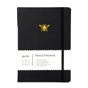 May include: Black dotted notebook with a gold bee emblem. The notebook has a black elastic band closure and a white label with the text "Dotted Notebook" and product details. Features include a vegan leather cover and 160 numbered pages.