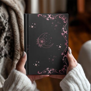 Op de afbeelding: Een zwart notitieboek met een roze bloemen- en hemels ontwerp. De omslag heeft een halve maan met een bloem, sterren en decoratieve hoekaccenten. Het notitieboek wordt vastgehouden door een persoon die een witte trui draagt.