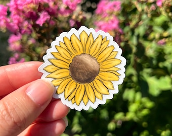 May include: A yellow sunflower sticker with a brown center. The sticker is held in a hand against a blurred background of pink and green flowers.