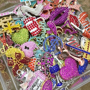 May include: A clear plastic container filled with a variety of colorful rhinestone charms. Charms include high heels, hearts, perfume bottles, and musical instruments. The charms are in shades of pink, red, blue, green, and gold.