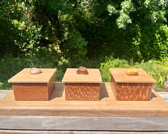 May include: Three small, wooden boxes with lids, each topped with a different-colored stone. The boxes are set on a rectangular wooden base. The boxes have a reddish-brown, textured pattern.