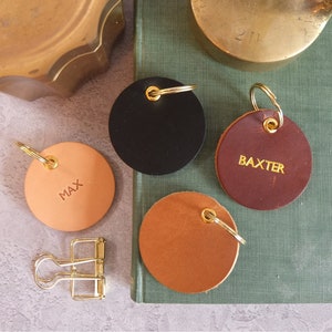 Può includere: Quattro portachiavi in ​​pelle con anelli dorati. I portachiavi sono rotondi e di diversi colori: nero, marrone, marrone chiaro e pesca. I portachiavi sono personalizzati con i nomi "MAX", "BAXTER" e due portachiavi vuoti.