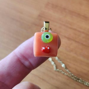 Puede incluir: Un colgante cuadrado de color naranja que presenta un solo ojo de color verde lima con una pupila negra y una pequeña pieza decorativa roja. El colgante está unido a una anilla de metal dorado y cuelga de una cadena a juego.