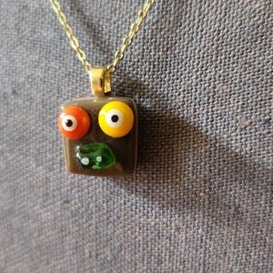Puede incluir: Un colgante cuadrado de vidrio marrón con un diseño de cara. El colgante tiene un ojo naranja, un ojo amarillo y un pequeño detalle de boca verde. Está suspendido de una fina cadena de eslabones de metal en tono dorado como collar.