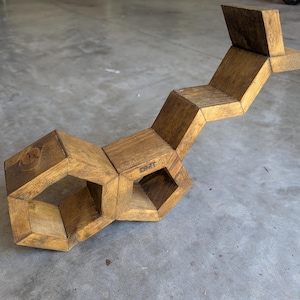 May include: A geometric wooden sculpture with a series of interconnected, angular blocks. The sculpture is a warm, medium brown colour and features open spaces. The word "DMT" is visible on one of the blocks. The sculpture is on a concrete surface.