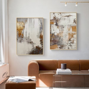 May include: Two abstract paintings with a white background and brown, gray, and gold accents. The paintings are framed in gold and hung on a white wall above a brown couch.