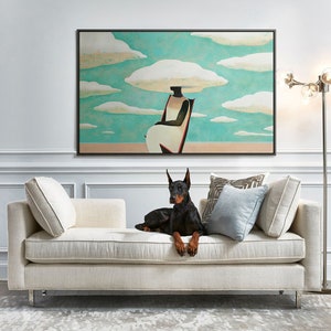 May include: A black Doberman Pinscher dog lays on a white linen sofa with a large abstract painting of a woman sitting on a chair with clouds above her head hanging on the wall behind the sofa.