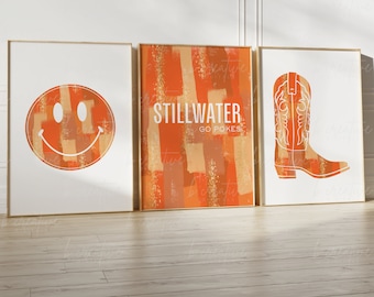 May include: Three framed prints with an orange and gold abstract design. The first print features a smiley face, the second print says "Stillwater Go Pokes", and the third print features a cowboy boot.