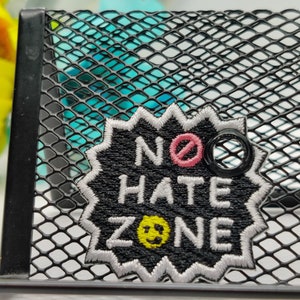 May include: Black embroidered patch with white stitching that says "NO HATE ZONE" with a pink circle and diagonal line through it and a yellow smiley face.