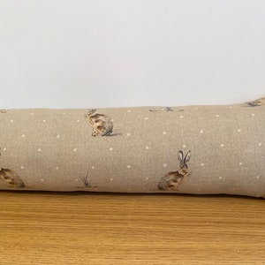 May include: A long, rectangular doorstop with a beige linen fabric cover. The fabric has a white polka dot pattern and a repeating design of brown rabbits.