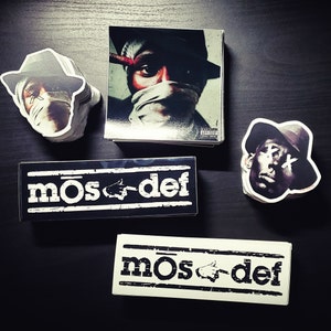 Könnte beinhalten: Drei schwarz-weiße Aufkleber mit dem Text "mOs def" in fetter, weißer Schrift. Die Aufkleber befinden sich auf einer dunklen Holzoberfläche.