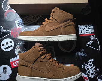 May include: A pair of brown suede high-top trainers with white soles and a Nike swoosh logo on the side. The trainers are sitting on a surface with various stickers.