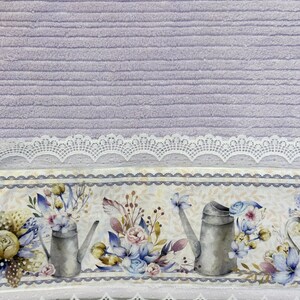Puede incluir: Toalla morada con un borde de encaje blanco y un estampado floral con regaderas y flores. El estampado es en tonos de azul, amarillo y verde.