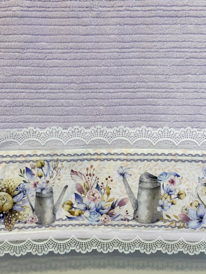 Puede incluir: Toalla morada con un borde de encaje blanco y un estampado floral con regaderas y flores. El estampado es en tonos de azul, amarillo y verde.