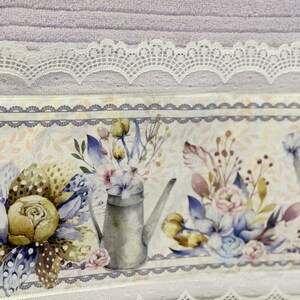 Puede incluir: Una toalla lavanda con un borde de encaje blanco y un estampado floral con una regadera y flores. La impresi&oacute;n es sobre un fondo blanco con un borde gris.