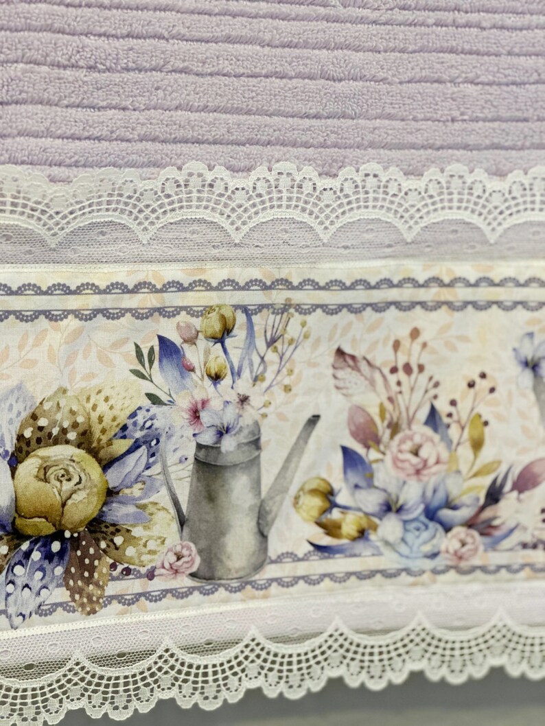 Puede incluir: Una toalla lavanda con un borde de encaje blanco y un estampado floral con una regadera y flores. La impresi&oacute;n es sobre un fondo blanco con un borde gris.