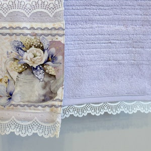 Puede incluir: Una toalla de mano lavanda con un dise&ntilde;o floral y un borde de encaje. El dise&ntilde;o floral presenta una regadera con flores y hojas. La toalla tiene una textura suave y absorbente.