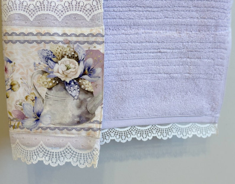 Puede incluir: Una toalla de mano lavanda con un dise&ntilde;o floral y un borde de encaje. El dise&ntilde;o floral presenta una regadera con flores y hojas. La toalla tiene una textura suave y absorbente.
