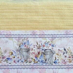 Puede incluir: Una toalla de cocina amarilla con un borde de estampado floral. El borde presenta un delicado dise&ntilde;o de encaje y dos regaderas con detalles florales.