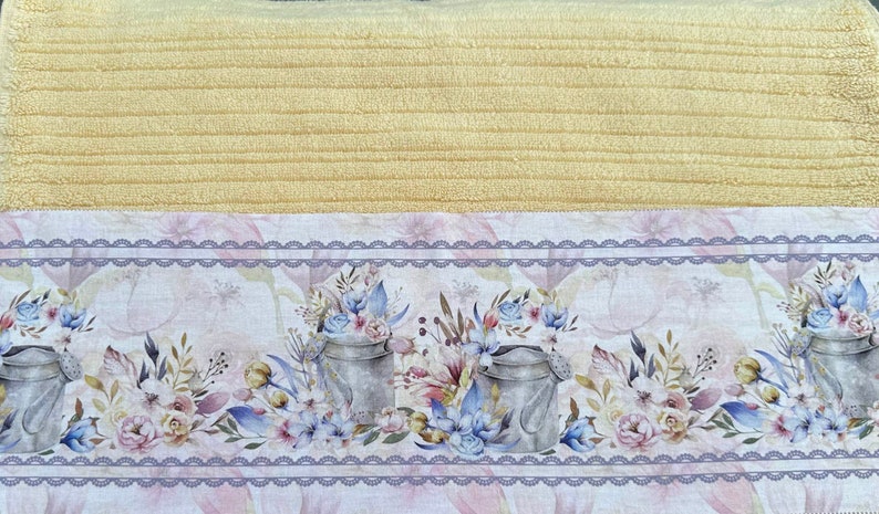 Puede incluir: Una toalla de cocina amarilla con un borde de estampado floral. El borde presenta un delicado dise&ntilde;o de encaje y dos regaderas con detalles florales.