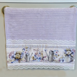 Puede incluir: Una toalla de mano lavanda con un borde de encaje blanco y un estampado floral con regaderas y flores.