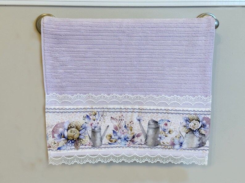 Puede incluir: Una toalla de mano lavanda con un borde de encaje blanco y un estampado floral con regaderas y flores.