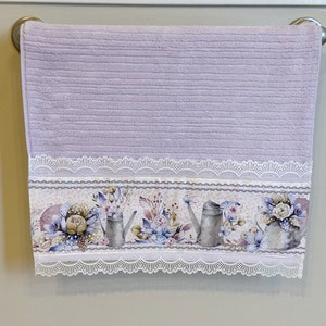 Puede incluir: Una toalla de mano lavanda con un borde de encaje blanco y un estampado floral con regaderas y flores.