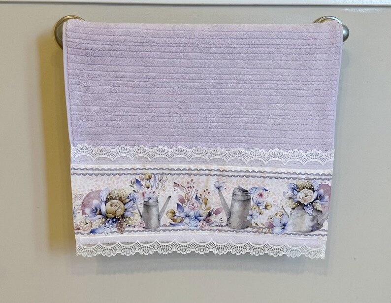 Puede incluir: Una toalla de mano lavanda con un borde de encaje blanco y un estampado floral con regaderas y flores.