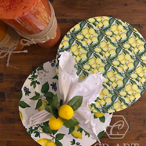 May include: Two round lemon-themed placemats with a white napkin and lemon accents. The placemats feature a green and yellow lemon design with white accents. The napkin is folded and has a lace trim. The placemats are on a wooden surface.