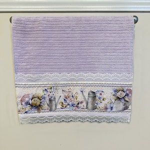 Puede incluir: Una toalla de mano lavanda con un borde de encaje blanco y un estampado floral con regaderas y flores.