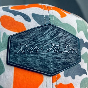 May include: A close-up of a camouflage-patterned baseball cap. The cap features a white background with orange, green, and gray shapes. A black, textured patch with the words "Ronin Fur Liner" is centered on the front.