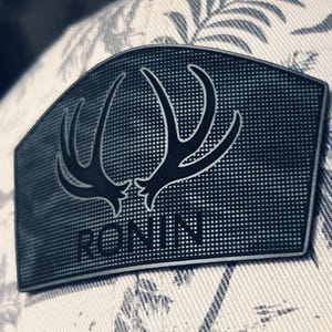 May include: A baseball cap with a white and grey floral pattern and a mesh back. It has a black patch with a deer antler design and the word "RONIN". The cap has a curved peak.