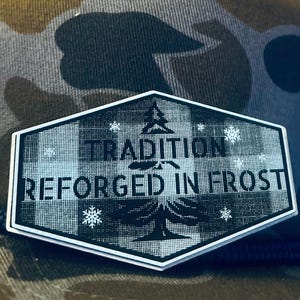 May include: A camouflage pattern baseball cap with a navy blue rope brim. The cap features a rectangular patch with the words "TRADITION REFORGED IN FROST" and a tree graphic.