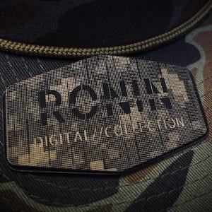 May include: A camouflage-patterned hat with a black brim and a decorative patch. The patch features the text "RONIN DIGITAL COLLECTION" in a pixelated design. The hat's colors include shades of green, brown, and black.