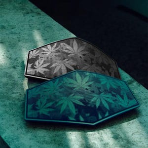 Puede incluir: Dos altavoces hexagonales con un patrón de hojas de cannabis. Un altavoz es negro y el otro es verde azulado. Los altavoces están sobre una superficie texturizada de color verde claro. Los altavoces tienen un diseño perforado.