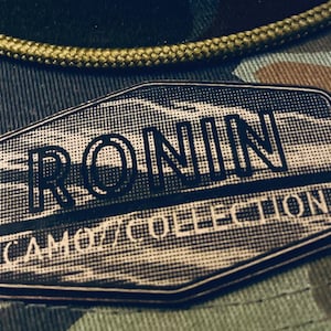 Könnte beinhalten: Nahaufnahme einer Camouflage-Baseballkappe mit einem Aufnäher mit der Aufschrift "RONIN CAMO COLLECTION". Die Kappe hat ein grün-braunes Camouflagemuster und ein grünes Seildetail.