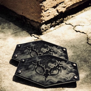 May include: Two black, hexagonal leather patches with a deer antler design. Each patch has four silver rivets and a textured surface. The patches are displayed on a concrete surface.