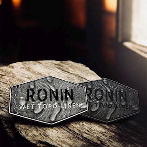 May include: Two hexagonal metal badges with the text "RONIN WET TOPO LINEN" in a dark, textured design. The badges are displayed on a piece of weathered wood, with a blurred background.