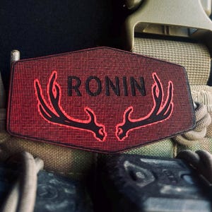 May include: A red patch with the word "RONIN" in black capital letters and a black antler design. The patch is in a hexagonal shape and is attached to a tactical vest. The background is dark.