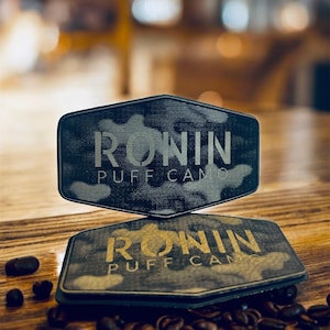 May include: Two black and gray patches with the text "RONIN PUFF CAMP" in a stylized font. The patches have a camouflage pattern and are displayed on a wooden surface with coffee beans scattered around.