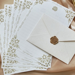 K&ouml;nnte beinhalten: Ein Stapel wei&szlig;es Briefpapier mit goldenen, verzierten R&auml;ndern und ein einzelner wei&szlig;er Umschlag mit einem goldenen Siegel aus Wachs. Das Papier tr&auml;gt den Aufdruck "Writing paper".