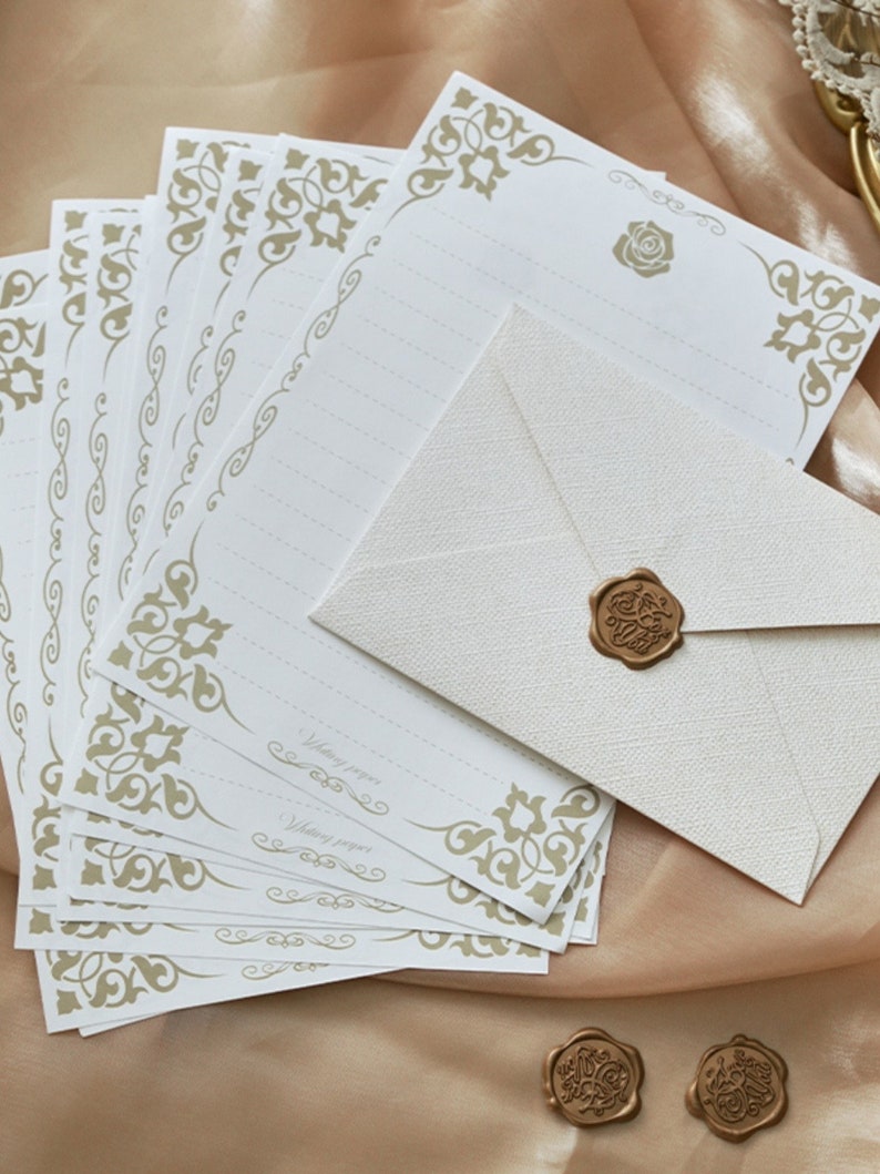 K&ouml;nnte beinhalten: Ein Stapel wei&szlig;es Briefpapier mit goldenen, verzierten R&auml;ndern und ein einzelner wei&szlig;er Umschlag mit einem goldenen Siegel aus Wachs. Das Papier tr&auml;gt den Aufdruck "Writing paper".