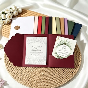 May include: A deep red tri-fold wedding invitation suite featuring a white embossed floral card and a separate save the date card with green leaf illustrations. The set includes a variety of colored envelopes and a gold wax seal on a white envelope.