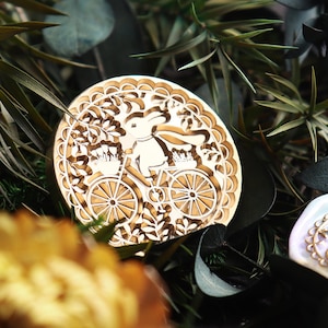 Pu&ograve; includere: Un timbro in metallo dorato con un design dettagliato di un coniglio che guida una bicicletta con fiori nel cestino. Il timbro &egrave; circondato da un bordo decorativo.