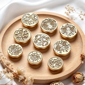 Könnte beinhalten: Eine Sammlung von neun runden Wachssiegelstempeln aus goldfarbenem Metall, die jeweils ein einzigartiges geprägtes Bienenmotiv mit Blumen- oder geometrischen Mustern zeigen und auf einem runden Holztablett mit glatter Oberfläche liegen.