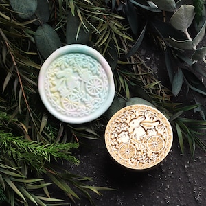 Pu&ograve; includere: Due timbri rotondi per sigilli di cera, uno con un design verde pastello, rosa e bianco di un coniglio che guida una bicicletta, e l'altro con un design dorato di un coniglio che guida una bicicletta.