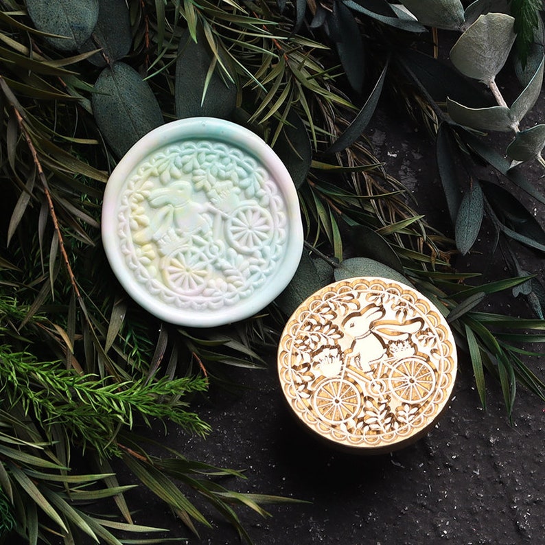 Pu&ograve; includere: Due timbri rotondi per sigilli di cera, uno con un design verde pastello, rosa e bianco di un coniglio che guida una bicicletta, e l'altro con un design dorato di un coniglio che guida una bicicletta.