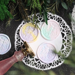 Pu&ograve; includere: Quattro sigilli di cera color pastello con un motivo floreale e un piccolo coniglio al centro. I sigilli sono su un pizzo bianco con un timbro di sigillo di cera dorato accanto.