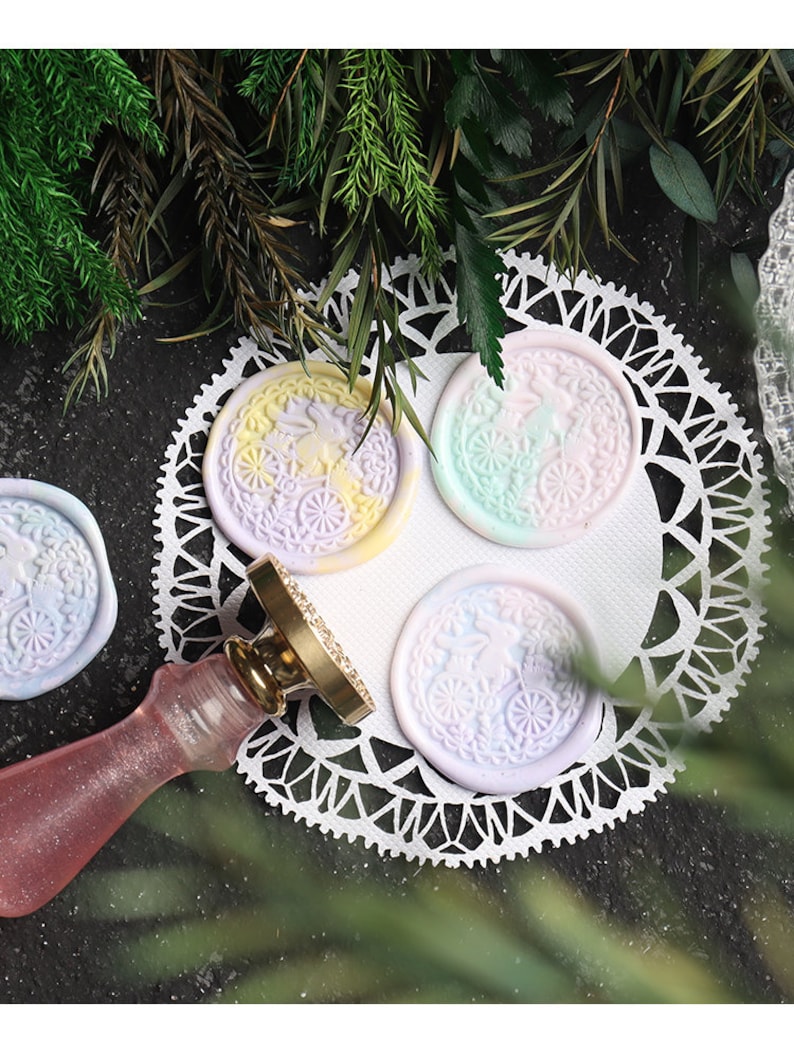 Pu&ograve; includere: Quattro sigilli di cera color pastello con un motivo floreale e un piccolo coniglio al centro. I sigilli sono su un pizzo bianco con un timbro di sigillo di cera dorato accanto.